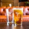 Laser-etched pint glass being filled with beer, with a second glass in background—great for team gifts, branded barware, or hospitality marketing.