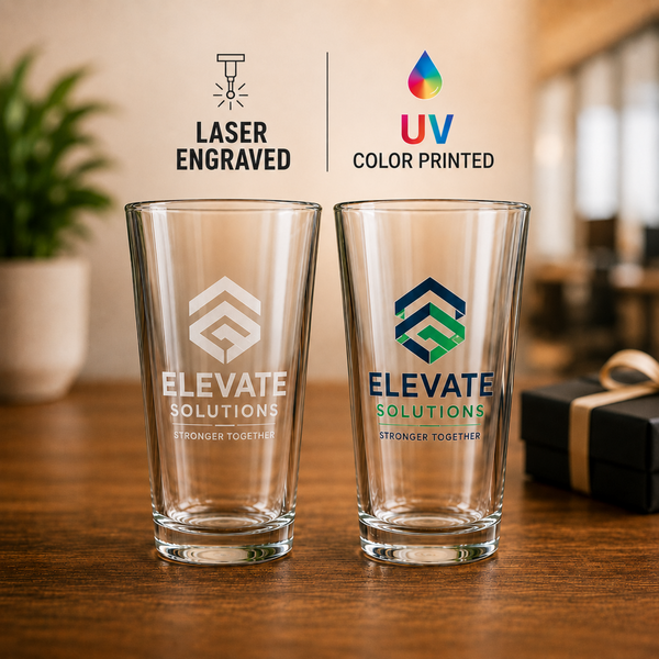 Two clear glass pint glasses on a wooden table, one with a white laser-engraved logo and the other with a blue and green UV color-printed logo. Above them, text reads 'LASER ENGRAVED' and 'UV COLOR PRINTED' with corresponding icons.