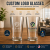 Two clear custom logo drinking glasses displayed on a wooden table, with promotional text above reading 'CUSTOM LOGO GLASSES' and feature icons for employee appreciation, client gifts, events & trade shows, and corporate giveaways. The glasses show the Elevate Solutions branding, and callout badges mention laser engraving and UV full color printing.