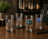 Four clear engraved drinking glasses displayed on a wooden table in a warm, blurred indoor setting. The glasses feature different corporate-style logos and text: 'ELEVATE SOLUTIONS', 'SUMMIT ADVISORS', 'PINNACLE FINANCIAL', and 'GLOBAL OUTREACH'.